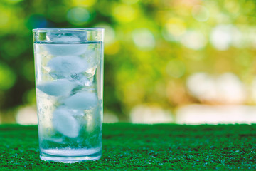 Cool water into a glass of ice with blurred natural green background and added color filter