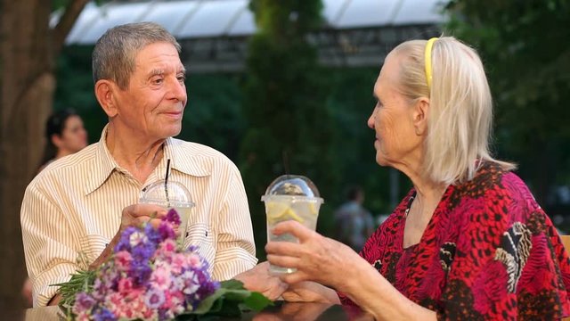 An elderly married couple sitting in a cafe outdoors with lemonade, slow motion.