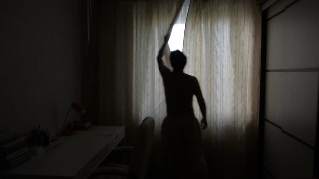 Man with a backlight opens the curtains on a sunny morning. The person opens the curtains and stretches the arms. Wide corner of the room