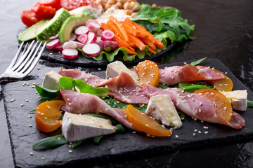 Two different salads om black stone slate. Vegan salad. Meat salad. Salad with ham jamon serrano, camembert, melon, arugula