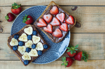 Plate with fruit toasts. Breakfast sandwiches with chocolate butter, banana slices, strawberry and blueberry. Heart shaped fruits. Sweet food. Fruit snack