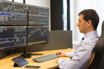 Stock broker trading online while accepting orders by phone. Trader's office with multiple computer screens full of index charts and data analyses.