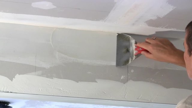 Homeowner uses putty knife to spread joint compound over seam of drywall between wall and ceiling.