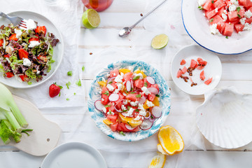 Variety of raw fresh summer salads with fruits and berries on white wooden background served with napkins and drinks. Summertime organic easy lifestyle concept. Flat lay with copy space.