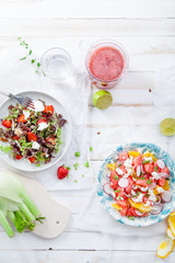 Variety of raw fresh summer salads with fruits and berries on white wooden background served with napkins and drinks. Summertime organic easy lifestyle concept. Flat lay with copy space.