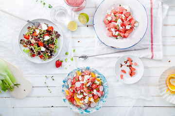 Variety of raw fresh summer salads with fruits and berries on white wooden background served with napkins and drinks. Summertime organic easy lifestyle concept. Flat lay with copy space.