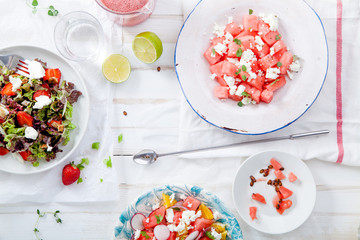 Variety of raw fresh summer salads with fruits and berries on white wooden background served with napkins and drinks. Summertime organic easy lifestyle concept. Flat lay with copy space.