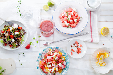 Variety of raw fresh summer salads with fruits and berries on white wooden background served with napkins and drinks. Summertime organic easy lifestyle concept. Flat lay with copy space.