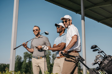 Three smiling men in sunglasses holding golf clubs outdoors - Powered by Adobe