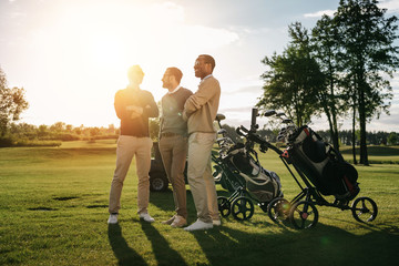 Three smiling men standing with crossed arms near golf clubs in bags