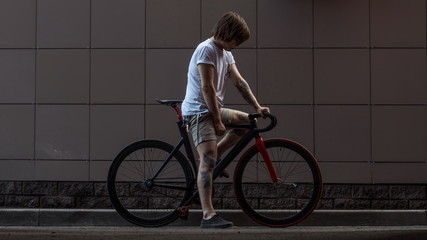 Tattooed biker hipster man in shorts standing against a grey wall next to a fixed gear bike