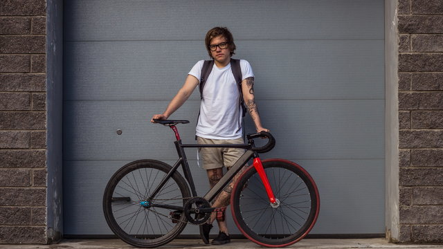 Tattooed biker hipster man in shorts standing next to a fixed gear bike against the background of gray rolling gates