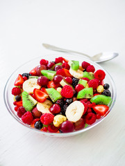 Fruit salad in glass plate with banana, kiwi and berries on white wooden background.
