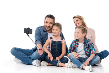 Fototapeta na wymiar smiling family taking selfie on smartphone while sitting on floor isolated on white