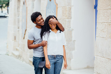 Obraz na płótnie Canvas Romantic lovely african american couple meeting on city street , man close her eyes and make surprise, dressed in jeans, summertime. Love couple and relationship concept