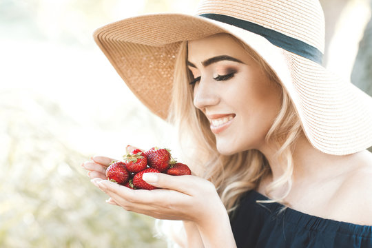 Smiling blonde woman holding strawberry and wearing straw hat outdoors in sunny day. Healthy eating. Summer season.