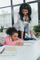 smiling mother looking at little daughter writing in notebook at workplace