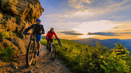 Naklejka na ściany i meble Mountain biking women and man riding on bikes at sunset mountains forest landscape. Couple cycling MTB enduro flow trail track. Outdoor sport activity.
