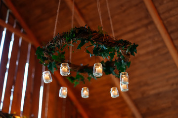 Small candles in jars hanging from green round garland on wooden background