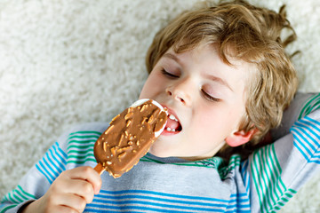 Little blond kid boy with curly hairs eating ice cream popsicle with chocolate at home