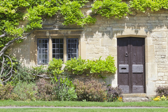 Dark brown old doors in traditional English stone house with climbing wisteria on the wall, flowers and herbs in a small front garden .