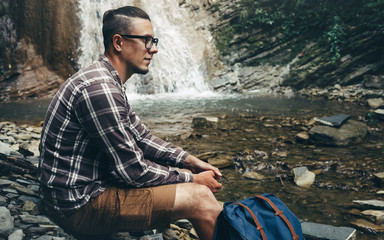 Traveler Sitting On The Rocks Near Waterfall. Adventure Travel Remote Relax Concept