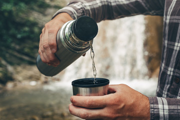 Unrecognizable Traveler Pouring Tea Or Coffee To Cup From Thermos On Waterfall Background. Tourism And Travel Concept
