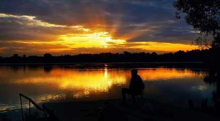 The silhouette of a seated fisherman on a beautiful background of a rising sun with rays in the dramatic clouds .
