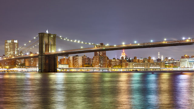 Panoramic photo of the Brooklyn Bridge with city lights reflected in East River at Night, New York City, USA.