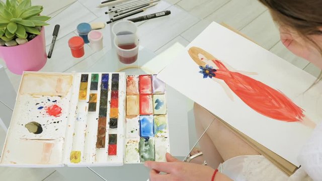 Female designer paints a sketch of her future red dress. Painting with watercolors on a white sheet of paper
