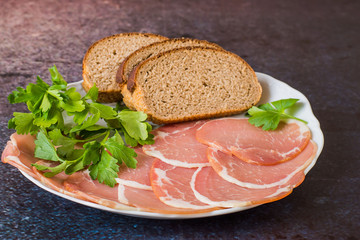 
 Fast snack. Smoked bacon, sliced thin slices, bread and fresh greens on a white porcelain dish on a dark background.