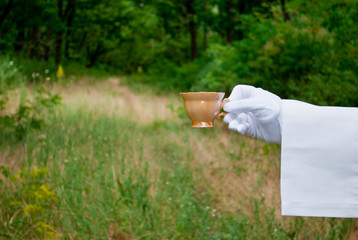 Fototapeta na wymiar The waiter's hand in a white glove and with a white napkin holds an empty pink beige small coffee cup on a blurred background of nature green bushes and trees