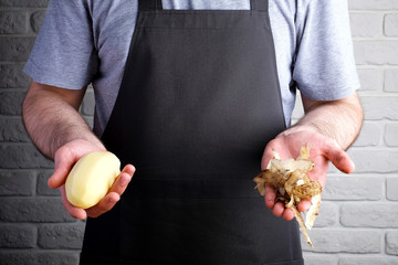 man in black apron holding peeled potatoes and potato skins in his hands, on background of brick wall