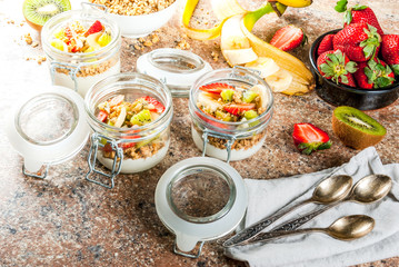 Healthy breakfast. Diet. Overnight oatmeal in a can, muesli. Yogurt with homemade granola and organic fruits - kiwi, banana, strawberry. On the stone table. With ingredients and spoons.