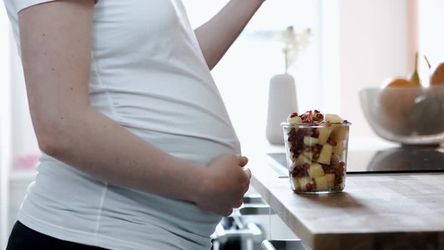 A pregnant woman eats pieces of pineapple and pomegranate from a glass cup.