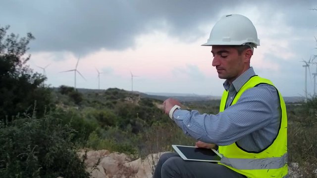 Employee of wind power working at a tablet on a background of windmills and looks at his watch