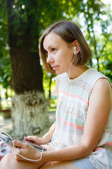 Young woman sitting on background of park and using headphones with tablet.