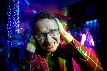 A man at a party in a suit covers his ears with hands from loud music. The man squeezed his eyes...
