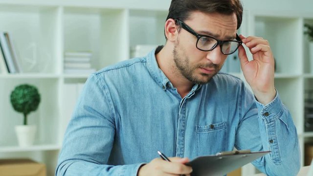 Young man in glasses checking final financial report from the laptop in the office background and feeling lost.