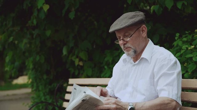 Handsome senior man reading the newspaper on the bench in park 4K