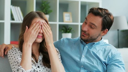Attractive man surprising his emotional girlfriend who clossing the eyes with a gift while they sitting on sofa at home.
