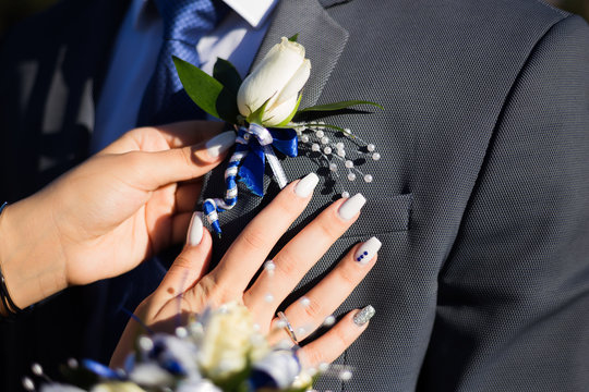 Womans hand with manicure is putting boutonniere on men suit. White rose with blue tape. Prom photo