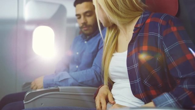 On a Plane Beautiful Blonde Female and Handsome Hispanic Male Fasten They're Seat Belts and Ready to Take off/ Landing. Sun Shines Through Airplane's Window. 4K UHD.