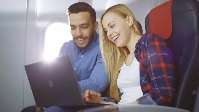 On a Board of Commercial Airplane Beautiful Young Blonde with Handsome Hispanic Male Watch Movies on a Laptop and Laugh. Sun Shines Through Aeroplane Window. 4K UHD.