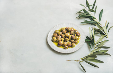 Pickled green Mediterranean olives in virgin olive oil on white ceramic plate and green olive tree...