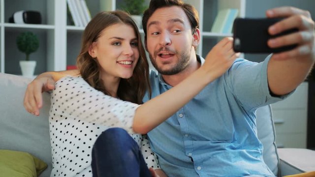 Happy young cuple in love making selfies on smart phone, making funny faces in a living room while sitting on the sofa at home.