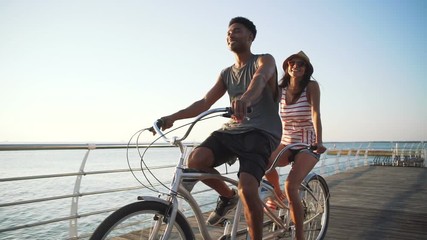 Portrait of a mixed race couple riding on tandem bicycle outdoors near the sea, slow motion - Powered by Adobe