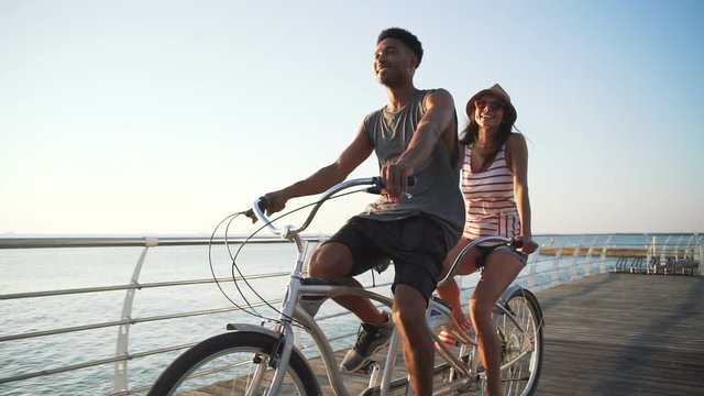 Portrait of a mixed race couple riding on tandem bicycle outdoors near the sea, slow motion
