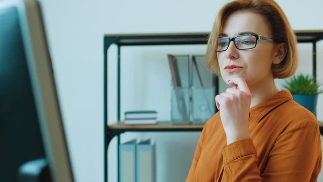 Young business woman in the glasses using laptop for video chating with business partners while siting at hte table in the office.