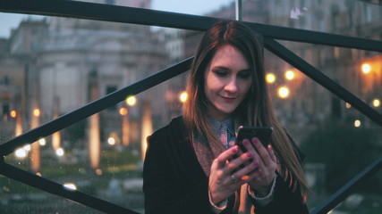 Young beautiful woman sitting in the city at evening and use smartphone. Girl texting with touchscreen keyboard.